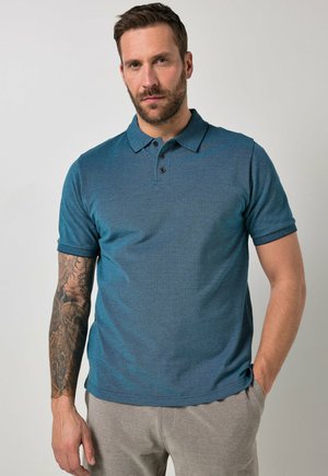 Mand med skæg iført en grønblå kortærmet polo og lysegrå bukser, med en ærmetatovering på venstre arm.