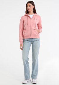 Felpa rosa con zip e coulisse, tasche frontali e polsini a costine, abbinata a jeans dritti azzurri e sneakers bianche.