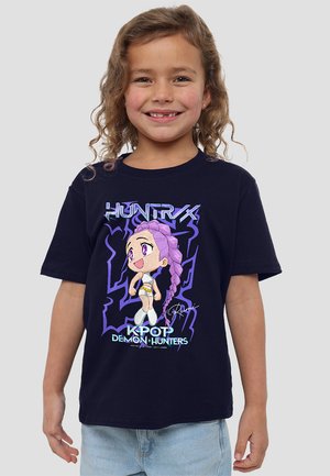 Ein junges Kind mit lockigem Haar trägt ein schwarzes T-Shirt, auf dem ein Anime-Mädchen mit lila Haaren und der Schrift "KPOP DEMON HUNTERS" auf der Vorderseite abgebildet ist.