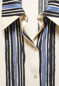 Gros plan sur une chemise crème avec des rayures verticales en traits de pinceau noirs et bleus et un col pointu, présentant trois boutons visibles.