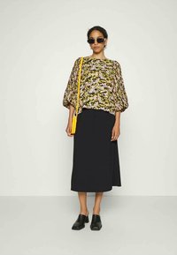 Mulher a usar uma blusa floral com mangas balão, saia midi preta, mules de salto preto, óculos de sol escuros e uma mala de ombro amarela.