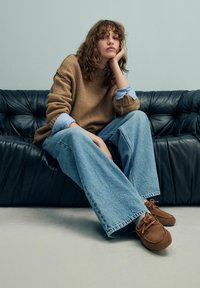 Bruine gebreide trui, lichtblauw shirt eronder, wijdvallende lichtblauwe jeans, bruine suède schoenen, zittend op een zwart leren bank.