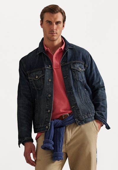 Σκούρο μπλε τζιν μπουφάν με τσέπες στο στήθος, φορεμένο πάνω από ροζ πόλο. Μπλε πλεκτό πουλόβερ δεμένο γύρω από τη μέση, σε συνδυασμό με μπεζ παντελόνι.