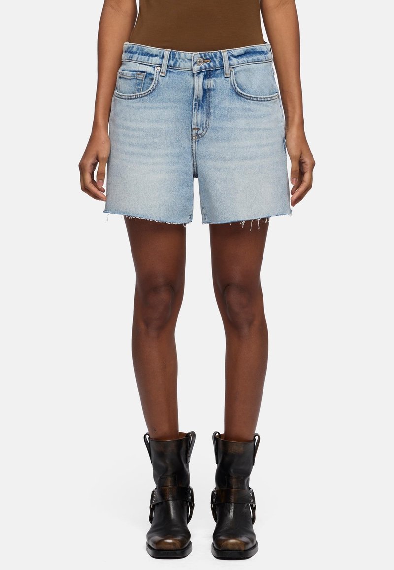 Persoon draagt lichtblauwe spijkershorts met rafelranden en zwarte leren laarzen tot halverwege de kuit, staand tegen een effen achtergrond.