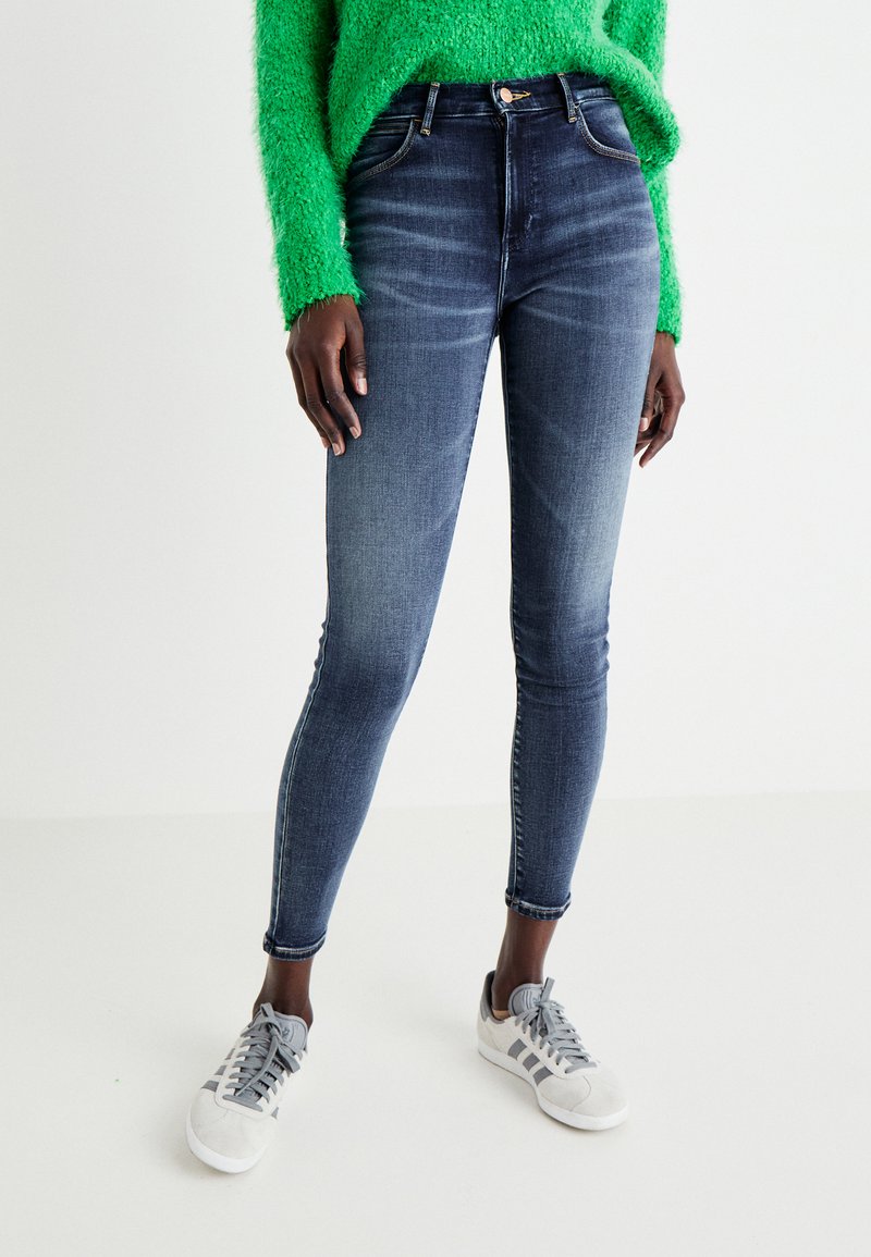 Tummansiniset kapeutetut farkut, joissa on korkea vyötärö ja tavallinen viiden taskun malli, sekä hienoinen kulahtanut pinta. Yhdistetty harmaisiin lenkkareihin.