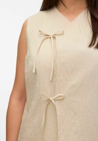 Beige mouwloze top met een V-hals, twee stoffen strikjes in de taille en een gestructureerde afwerking voor een lichte, ademende uitstraling.