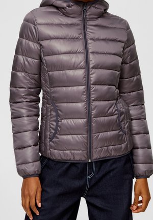 Osoba nosící šedou prošívanou bundu na zip s kapucí a tmavé kalhoty na jednoduchém světlém pozadí.