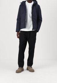 Marineblå hættejakke, hvid sweatshirt, sorte cargo-bukser og beige støvler. Jakken har lynlås og et strømlinet design.