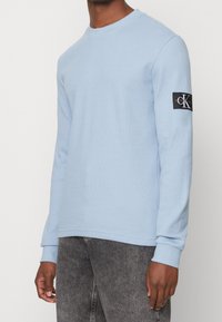 Muž v svetle modrém svetr s dlouhým rukávem a černou nášivkou loga Calvin Klein na levém rukávu, v kombinaci se světlými černými džíny.