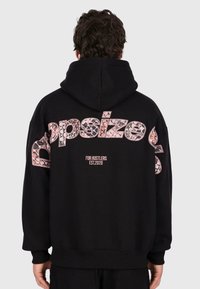 Man wearing black hoodie with large pink patterned "Dopsize" text and smaller "For Hustlers Est. 2020" text on back, standing facing away.