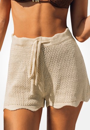 Mujer que lleva unos pantalones cortos beige de punto con cintura de cordón y un top de bikini marrón con textura, mostrada desde la mitad del torso hasta la mitad del muslo.