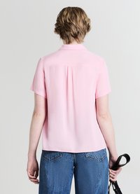 Blouse à manches courtes, rose clair, coupe droite, pli dos et col, fabriquée dans un tissu lisse. Portée avec un jean bleu taille haute.