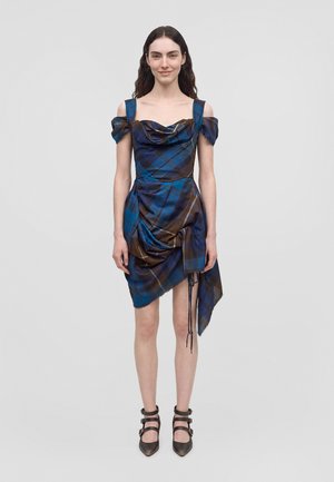 Un mannequin se tient devant un fond gris clair, vêtu d’une robe à carreaux bleus et bruns, épaules dénudées et panneaux drapés, ainsi que de chaussures noires à talons et à brides.