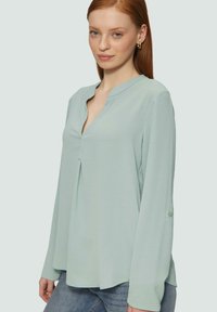 Blouse verte claire à manches longues avec un poignet à boutons, décolleté en V, détails de plis et tissu doux et texturé, portée avec un jean bleu.