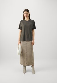 T-shirt à manches courtes gris foncé avec un motif texturé, associé à une longue jupe beige. Le modèle tient un sac à main blanc et porte des bottines grises claires.