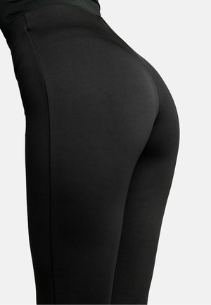Közeli kép egy személy szűk fekete leggingséről, amelyen a testhez simuló anyag látható a csípő és a comb felső része körül, egyszerű háttér előtt.