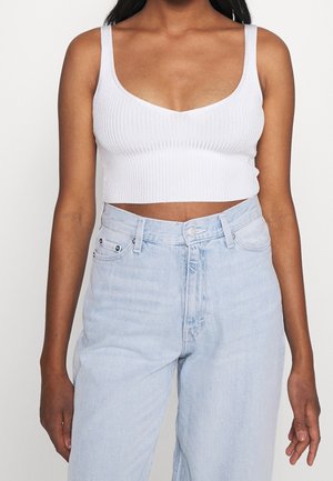 Femme portant un débardeur court blanc côtelé et un jean bleu clair taille haute sur un fond uni.