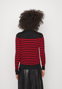 Maglione nero con strisce orizzontali rosse, caratterizzato da un design aderente e polsini a costine. Texture morbida con scollatura rotonda, indossato con una gonna nera.