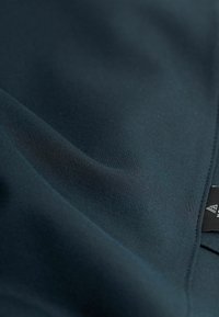 Tessuto verde scuro con una superficie liscia e opaca, caratterizzato da una leggera elasticità e un'etichetta con design geometrico. Bordo ripiegato visibile.
