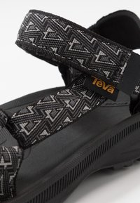 Sandaler med stropper som har et mønstret sort og grått tekstiloverdel, med en teksturert svart gummisåle og et fremtredende Teva-logo-merke.