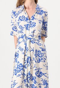 Modrá květinová košilová šaty s krátkými rukávy, knoflíkovým předním zapínáním a zavazovacím pasem. Vyrobené z lehkého, hladkého materiálu.