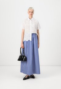 White eyelet blouse with a scalloped hem, paired with a blue and white striped maxi skirt. Model holds a small black bag and wears flat shoes.