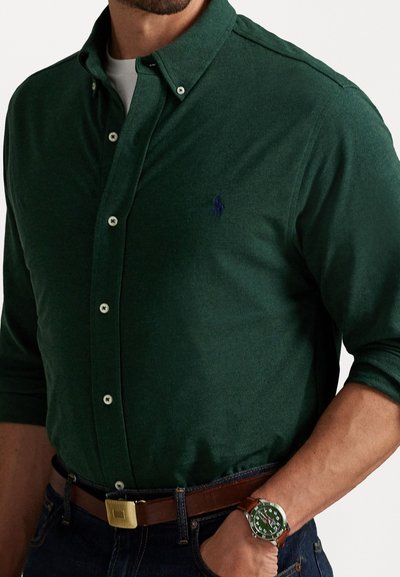 Mann med mørkegrønt skjorte med knapper, brun lærbelte, mørke jeans og grønne ansikt med håndleddsklokke med brun stropp, hånd i lomma.