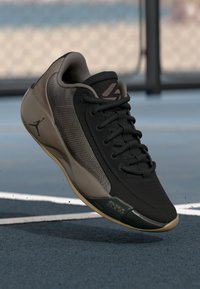 Čierne a béžové basketbalové tenisky s tkaničkami, so súmou Air Zoom a logom Jumpman, ktoré sú zobrazené nad modrým ihriskom s bielymi čiarami.