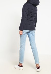 Mørkeblå pufferjakke med hætte, quiltet design og elastiske manchetter; parret med lyseblå jeans og hvide sneakers med maroon accenter.