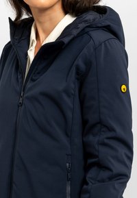 Marineblauwe gewatteerde jas met een ritssluiting, voorzien van een geel logo op de mouw, gemaakt van glad, licht gestructureerd stof.