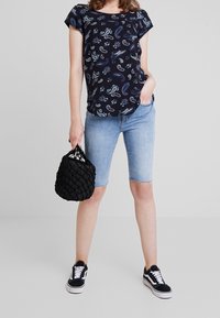 Top de manga corta en color navy con estampado paisley, combinado con pantalones cortos de mezclilla claros. Los accesorios incluyen un bolso negro trenzado y zapatillas negras.