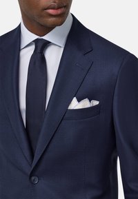 Veste de costume bleu marine en tissu texturé, fermeture à un bouton, et une poche poitrine avec un mouchoir en papier blanc plié. Chemise habillée blanche et cravate foncée.