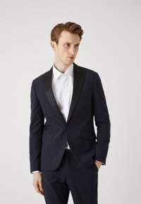 Abito blu navy con revers in raso nero, abbinato a una camicia bianca. Tessuto morbido, vestibilità slim con tasche laterali e bottoni in vista.