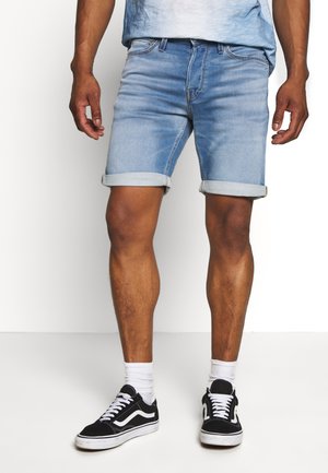 Shorts de mezclilla azul con puños enrollados, con un diseño clásico de cinco bolsillos. Combinados con zapatillas negras y calcetines blancos.