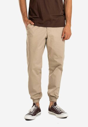 Mann trägt beige Jogger-Hose mit elastischen Bündchen, ein braunes Hemd und dunkelbraune Low-Top-Sneaker mit weißen Schnürsenkeln.