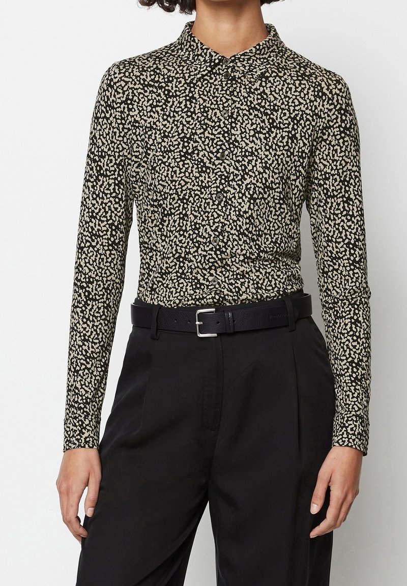 Femme portant une chemise à manches longues noire et beige à boutons avec des taches, rentrée dans un pantalon noir taille haute avec une ceinture noire.