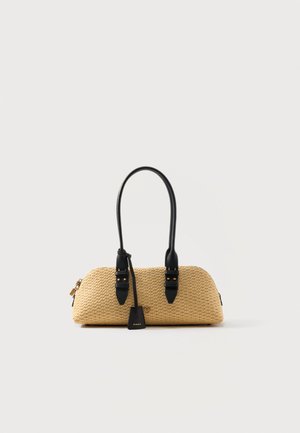Bolso pequeño tejido de color beige con asas largas negras, detalles de cuero negro y una pequeña etiqueta con la marca colgando de una de las asas.
