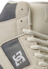 Beige och grå sneakers i mocka och meshmaterial, med grå accenter, vita snörningar och en framträdande logotyp på sidan.