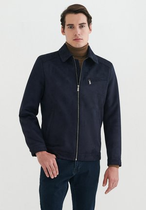 Mørkeblå faux suede jakke med lynlåsfortil, spids krave og en brystlomme. Har lange ærmer og en glat tekstur.