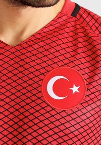 Maillot de sport rouge avec un motif en losanges texturé, un design en v, et un patch circulaire avec un emblème blanc en croissant et étoile.