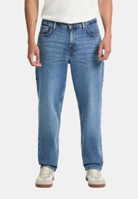 Jeans in denim azzurro chiaro con una vestibilità rilassata, design a cinque tasche e cuciture visibili, abbinati a sneakers beige e bianche.