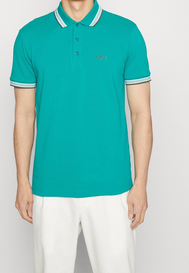 Homme portant un polo turquoise avec des bordures blanches et grises, logo BOSS sur la poitrine et pantalon blanc, debout devant un fond uni.