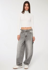 Młoda kobieta w białej, długiej koszulce z krótkimi rękawami i zamkiem, luźnych, wyblakłych szarych dżinsach i białych trampkach, stojąca na jednolitym tle.