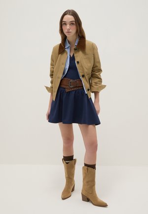 Vrouw draagt een beige jas met corduroy kraag, blauwe jurk, brede bruine gespikkelde riem en beige laarzen tot halverwege de kuit met donkere sokken tegen een effen achtergrond.