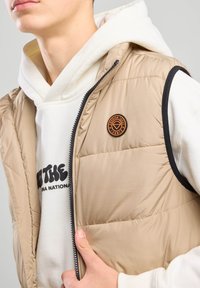 Beige tilattu liivi, jossa on vetoketju ja mustat yksityiskohdat. Siinä on pyöreä oranssi logo-täplä. Kerroksena valkoisen hupparin päällä, jossa on mustaa tekstiä.