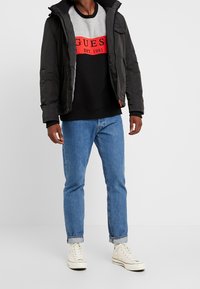 Chaqueta de plumas negra con cremallera, sudadera gris y negra con logo rojo de "GUESS", vaqueros azules y zapatillas blancas con ojales.