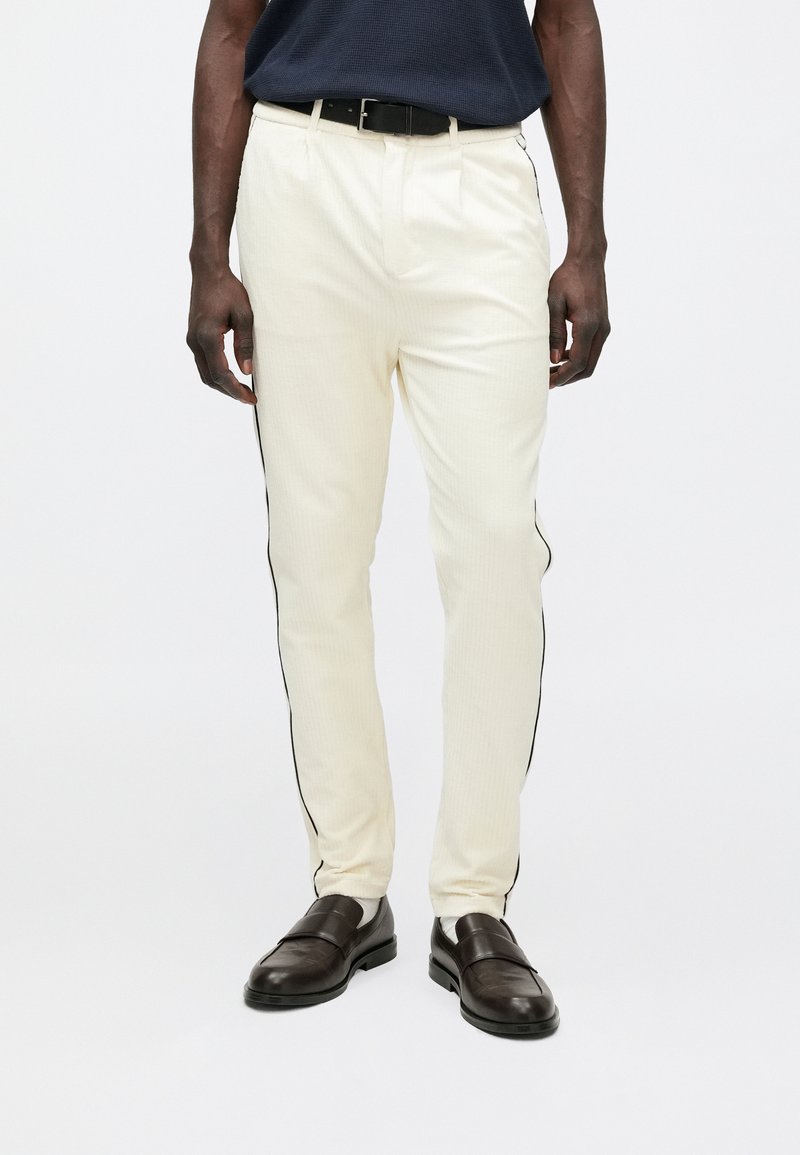 Kremowe spodnie o teksturowanej powierzchni, z czarnymi bocznymi paskami, w połączeniu z ciemnobrązowymi butami. Standardowy krój z paskiem.
