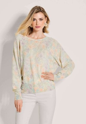 Frau mit blondem Haar trägt einen pastellfarbenen Blumenpullover und weiße Hose, posiert mit einer Hand in der Hüfte vor einfarbigem Hintergrund.