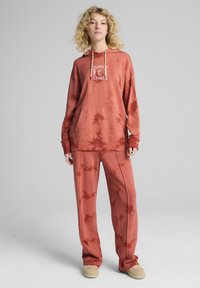 Hrdzavý tie-dye sweatshirt s prednou kapsou a kapucňou na šnúrku, doplnený o voľne strihané nohavice a béžovú obuv.
