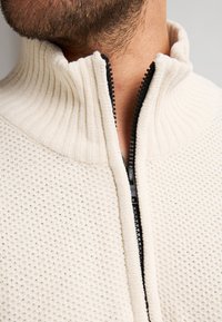 Maglione zip-up di colore crema con colletto a coste e tessuto testurizzato. Presenta una cerniera nera e un motivo a pois su tutta la superficie.
